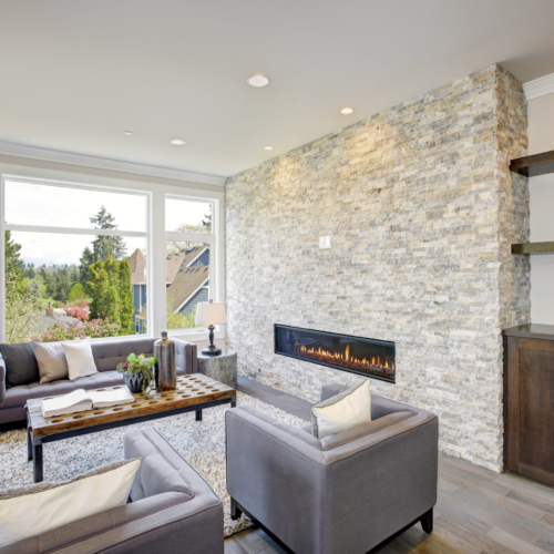 Modern great room features a floor to ceiling stone fireplace, gray tufted sofa paired with two gray armchairs over fluffy rug. Northwest, USA